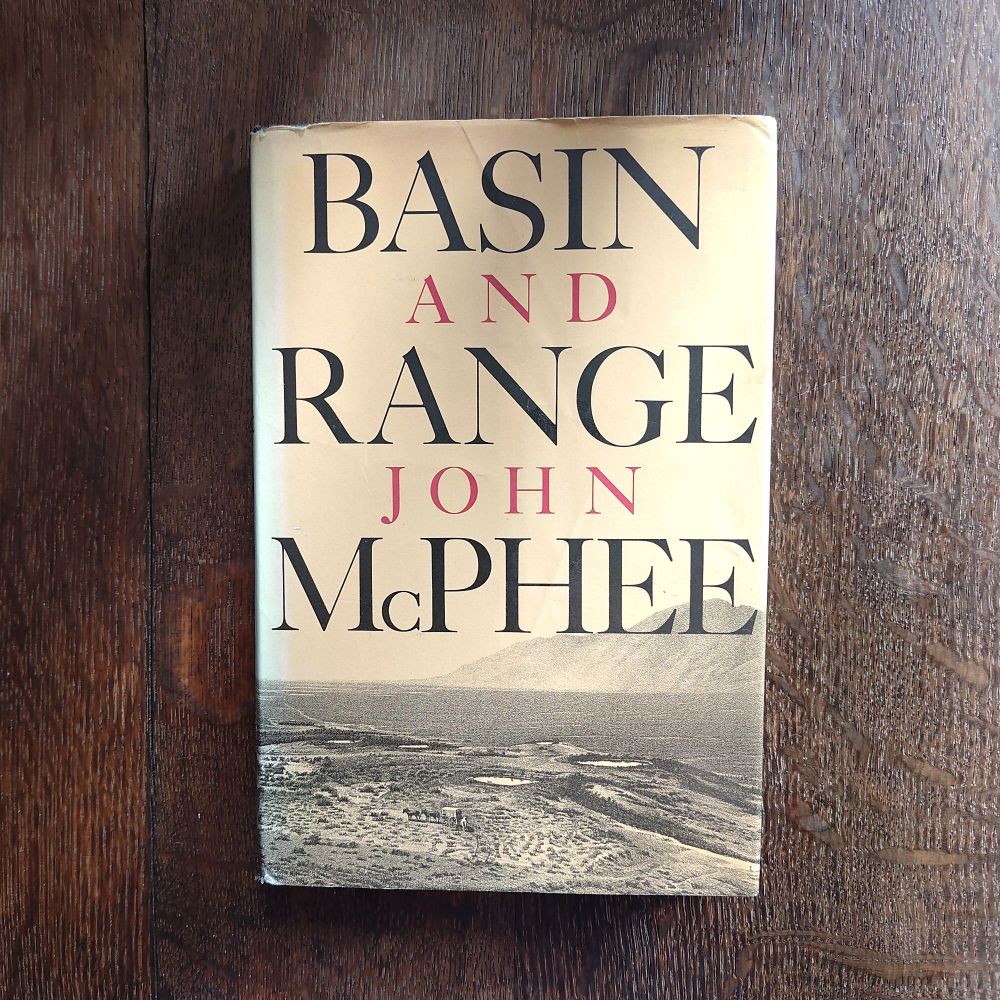 Book cover: Big, black main lettering, red sub lettering against a sepia photograph of a flat plain with small volcanic features in foreground, large mountain face in background. Basin and Range, John McPhee 
