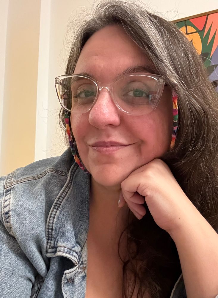 Selfie. Sou uma mulher branca, com cabelos compridos castanhos grisalhos. Estou com uma jaqueta jeans e óculos de armação transparente e cordinha colorida. Estou sorrindo levemente e apoiando o rosto na mão esquerda. 