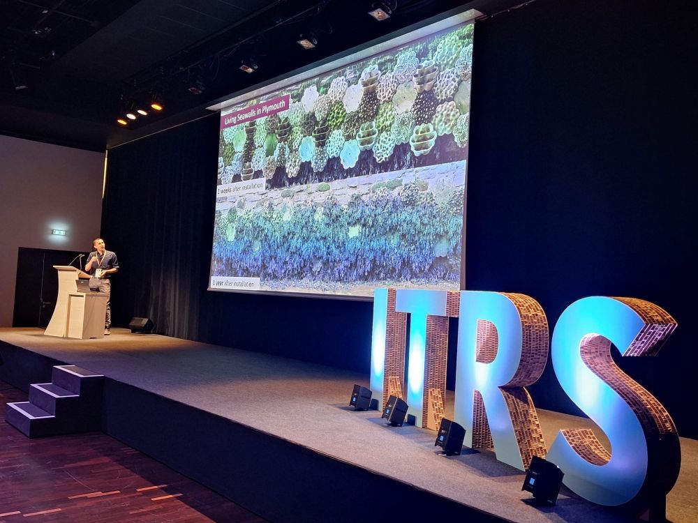 A speaker at a podium presenting with a large screen showing ITRS