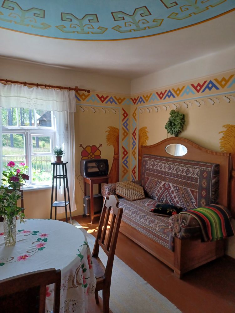 Eine hell und freundlich eingerichte Wohnstube. Vorne Stühle um einen runden Tisch, auf dem Tisch eine weiße Tischdecke mit wohldosierten bunten Stickereien und ein Blumenstrauß. Rechts an der Wand eine Holzbank mit hoher Lehne, darauf ein gemustertes Polster (oder ein Teppich?) und Kissen. Über dem Sofa hängt ein grüner Kranz an der Wand. Im Hintergrund steht eine Topfpflanze auf einem kleinen hölzernen Podest, daneben ein altes Radio (?) auf einem kleinen Holzregal. Wand und Zimmerdecke sind mit farbigen Zierstreifen bemalt. Durch das Fenster im Jintergrund scheint die Sonne.