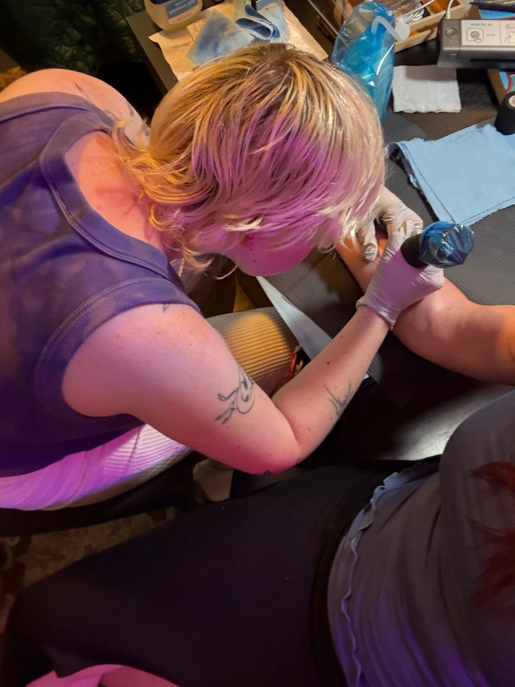 A blonde tattooed person tattooing the forearm of a woman