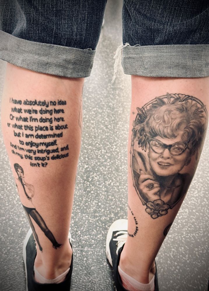 Photo of the back of a woman’s calves. On the right calf is a black and gray tattoo of Eileen Brennan as Mrs. Peacock in the 1980’s movie Clue. On the left calf is a tattoo of one of Mrs Peacock’s lines “I have absolutely no idea what we're doing here, or what l’m doing here, or what this place is about. But I am determined to enjoy myself and I’m very intrigued and oh my this soup's delicious isn't it?”