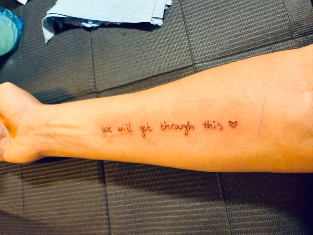 A tattoo on my forearm which reads “we will get through this ❤️” in brown ink