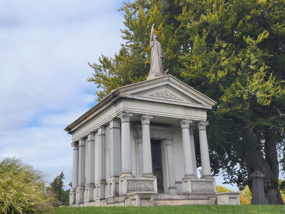 A Greek-temple style mausoleum set amongst trees. A figure stands atop with one arm raised skyward. 