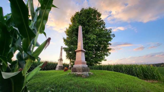 A rise of grassy land where three columnar gravestones stand alongside a tree.
The grave site is found amongst a cornfield. It is sunset and the sky is pale blue with whisps of cloud.