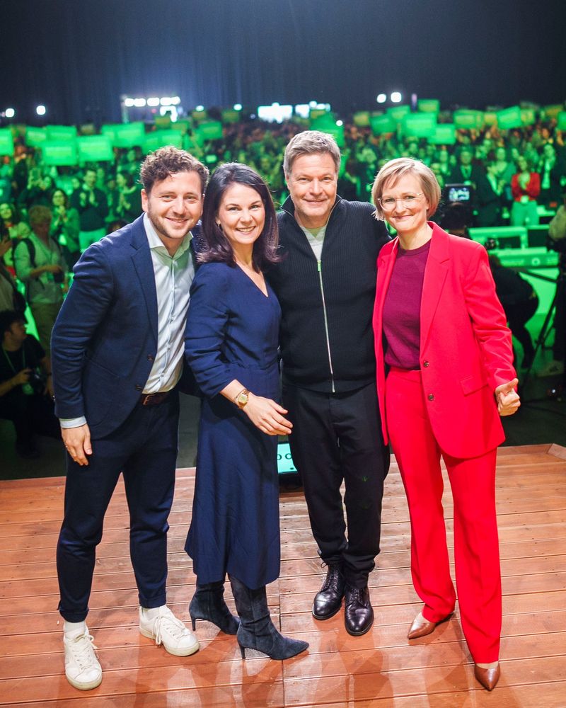 Felix Banaszak, Annalena Baerbock,Robert Habeck und Franziska Brantner posieren lächelnd auf einer Bühne, Publikum mit grünen Schildern im Hintergrund.