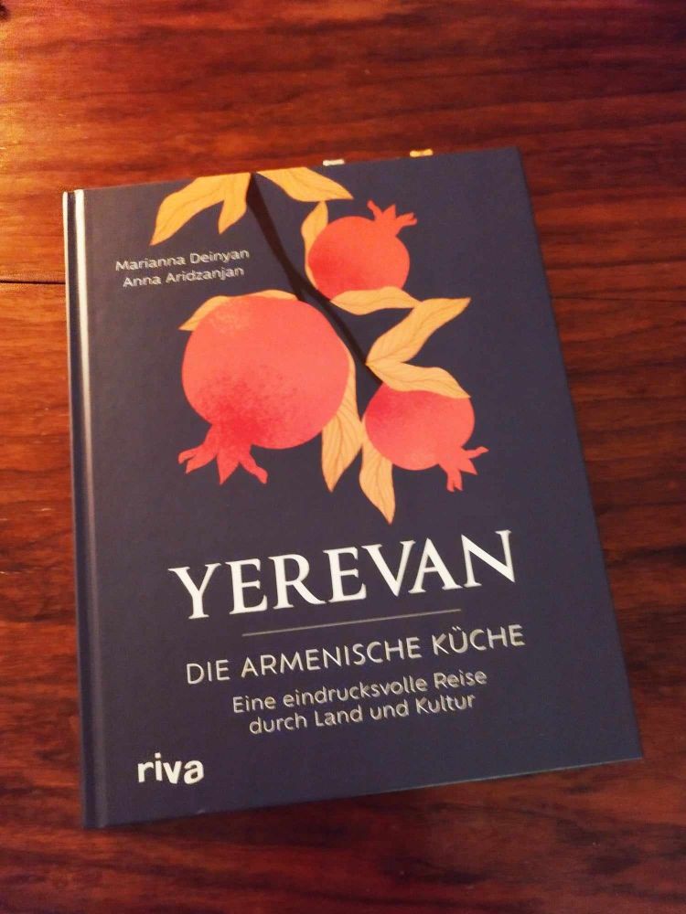 Yerevan Kochbuch zugeklappt auf Nussholztisch. Weiße Schrift auf dunkelblauem Untergrund plus popartige, orange-rote Illustration eines Granatapfelstrauchs.