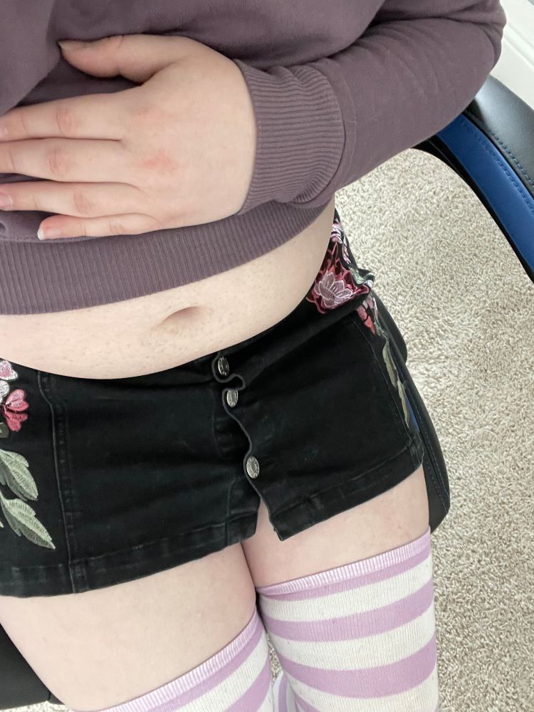 Me sitting in a chair, wearing a cute outfit, with a purple thigh highs, denim skirt, crop top, and exposed tummy, with my hand resting above my belly
