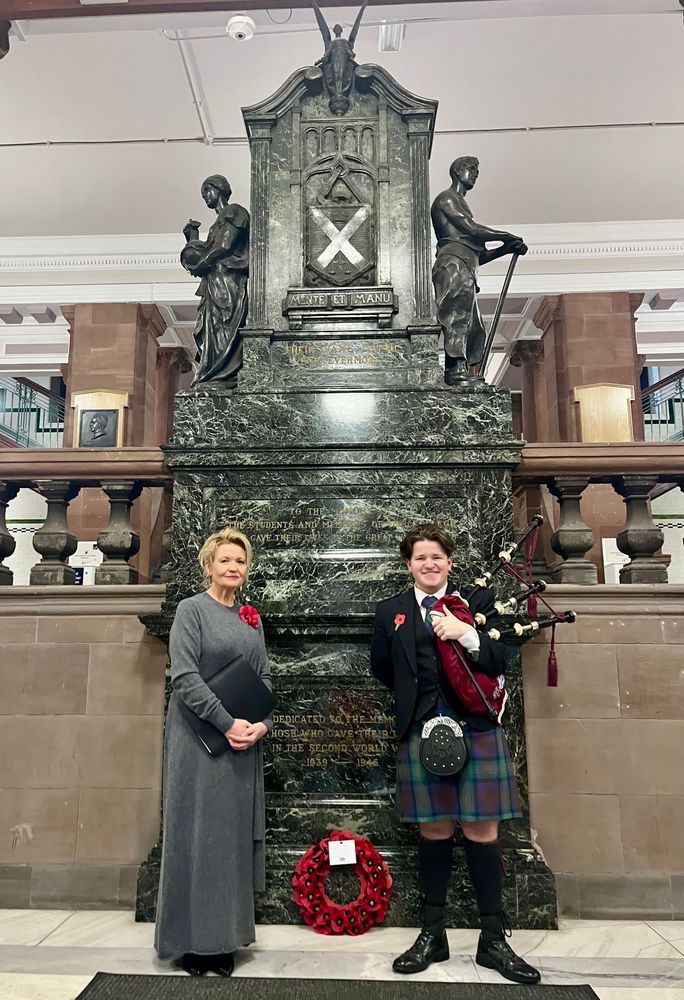 The photo shows Jennifer Buchan on the left in a long grey dress wearing a poppy, she is standing in front of a war memorial at the University of Strathclyde next to a piper in a kilt and holding bagpipes. A poppy wreath sits between them. 