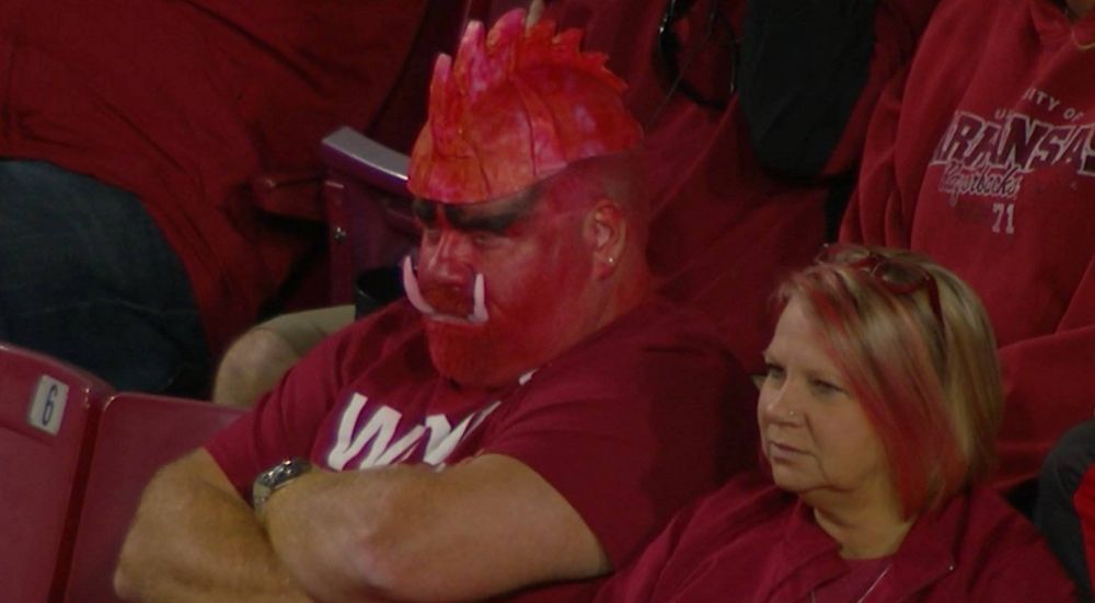 A Hog fan looking very upset. His arms are crossed. He is wearing red facepaint with white tusks and a cap made to look like a razorback mohawk.