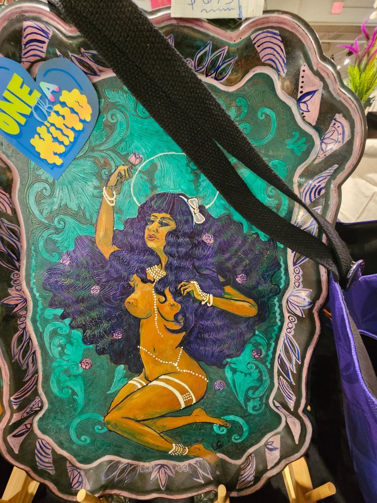 A nude brown woman with long curly black hair, decorated in gold chains, holding a rose is painted in acrylic on a solid silver serving tray. 