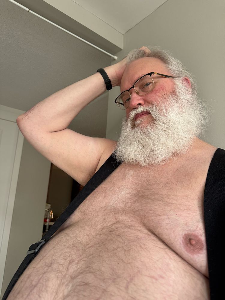 Bearish white haired man with a mischievous smile in suspenders, with his right hand on his head, ruffling his hair.