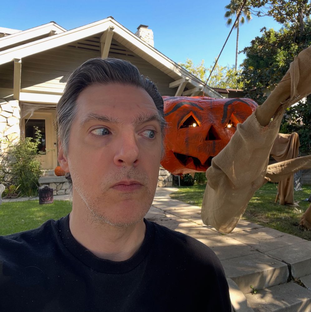 A man stands outside a house decorated for Halloween. He is looking behind himself. Behind him is a very large, scary, pumpkin spider guy with lights in their eyes.