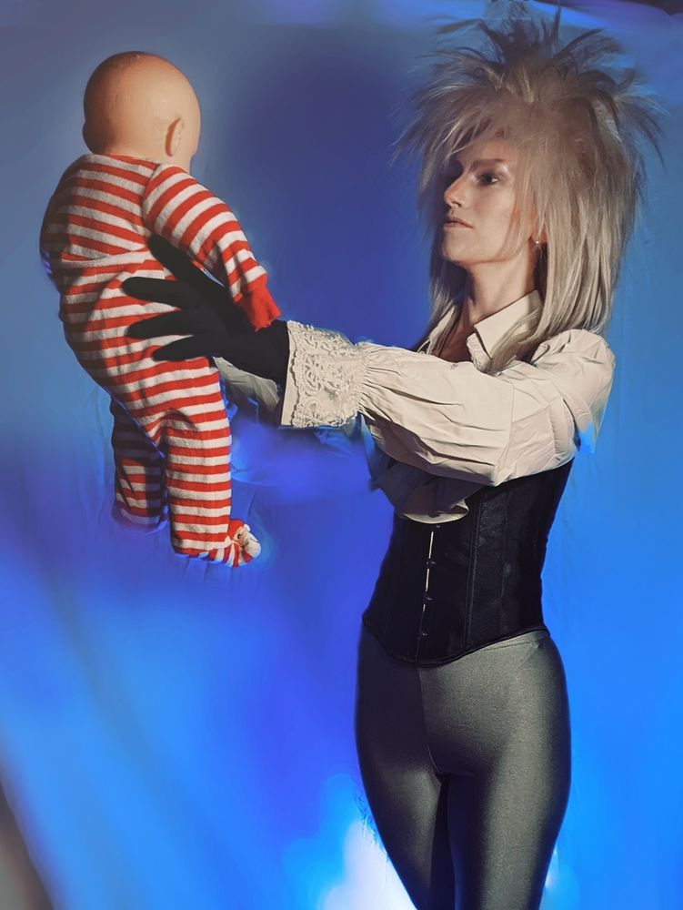 Cosplayer dressed as Jareth the goblin king from Jim Henson's Labyrinth against a blue backdrop, holding a baby doll in a striped onesie.
