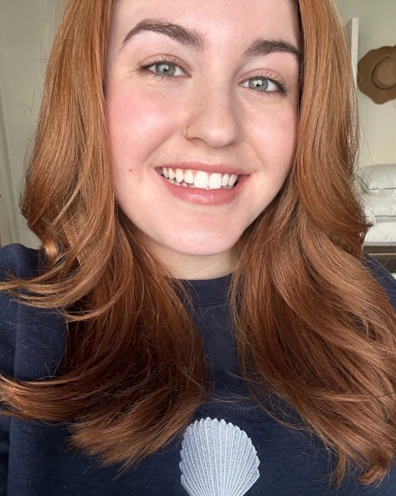 Smiling woman with auburn hair, green eyes, and a nose ring, wearing a navy sweatshirt with a seashell design.