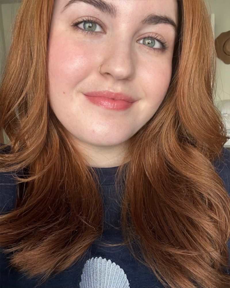 Young woman with auburn hair, green eyes, and a nose ring, wearing a navy sweatshirt with a seashell design, smiling softly.