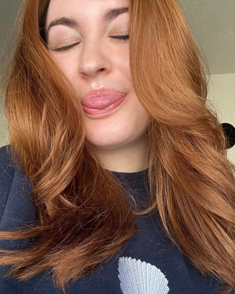 Young woman with auburn hair and a nose ring sticking out her tongue with closed eyes, wearing a navy sweatshirt with a seashell design.