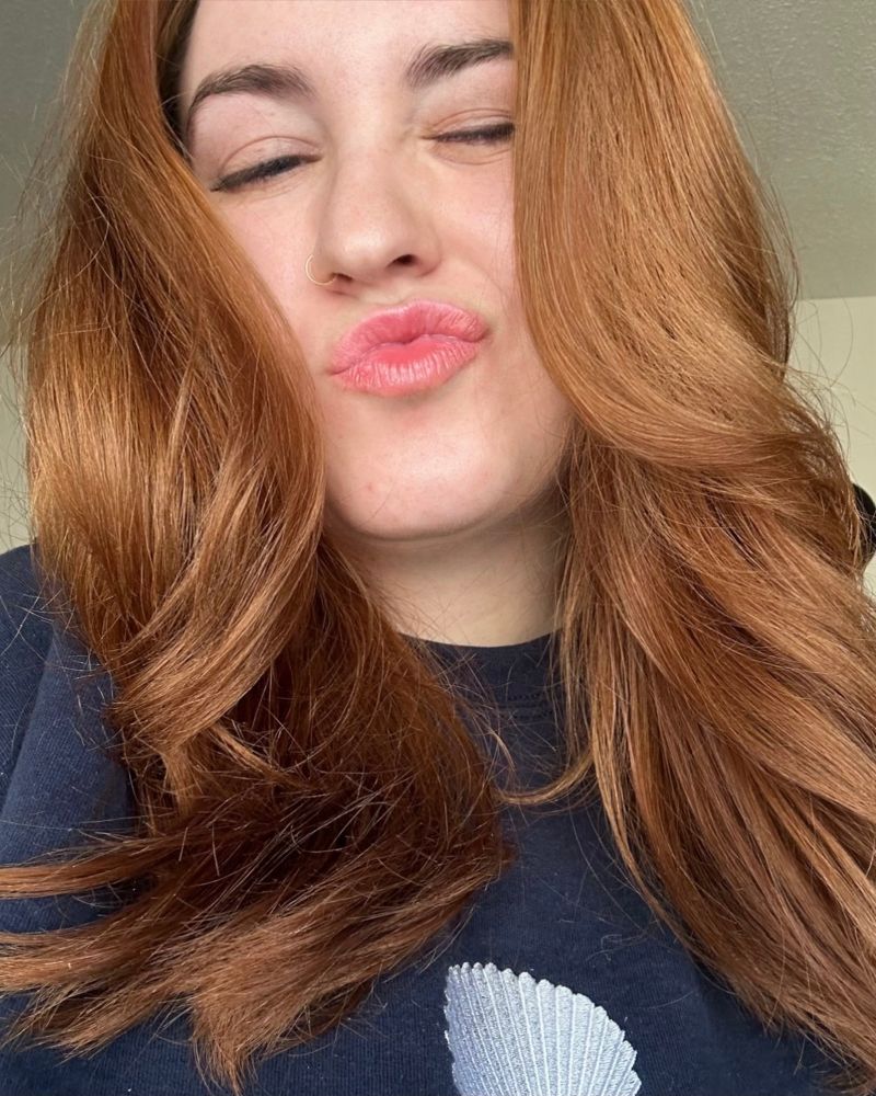 Young woman with auburn hair and a nose ring blowing a kiss with closed eyes, wearing a navy sweatshirt with a seashell design.”
