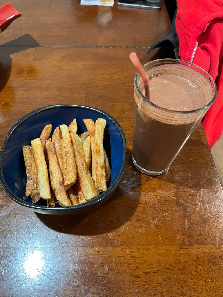 On a brown table sits a blue bowl that’s filled with fries. Next is a glass filled with a chocolate milkshake and a pink straw sticking out. On the top right corner is a part of a red hoodie resting on a chair.