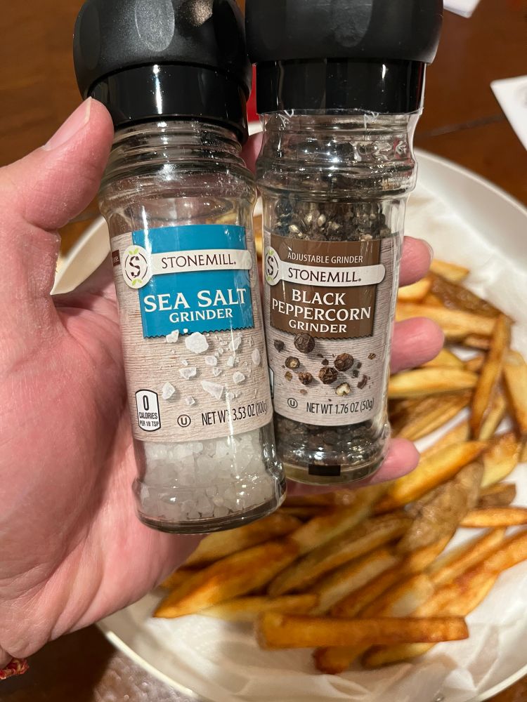 A hand is holding two containers. The one on the left is a glass container of sea salt with a built-in grinder, it’s a white label with blue accents. The one on the right is a glass container for black peppercorn with a built-in grinder, the label is white with brown accents. They are being held over a pan that houses some fries.