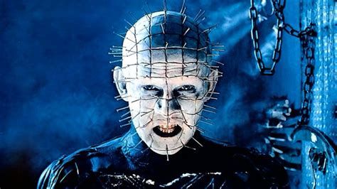 The Hell Priest (aka Pinhead) from the movie Hellraiser. It’s a being with nails evenly embedded in its head with the flat ends sticking out. He’s dressed in black leather and standing in front of some hanging chains. 