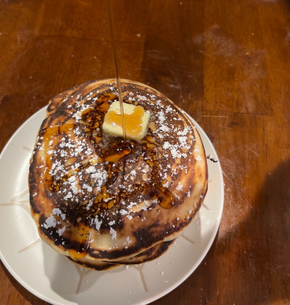 A stack of pancakes topped with powdered sugar, butter, and with syrup being poured on them. They are sitting on a white plate with a spiderweb design.