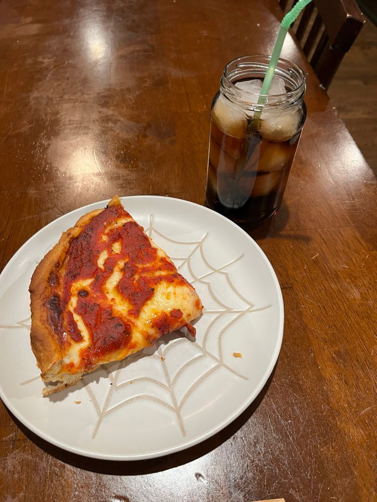 A slice of deep dish pizza on a white plate with a spiderweb pattern. In the top right of the plate is a glass of soda with ice and a green plastic straw sticking out of the top. They are sitting in a brown table.