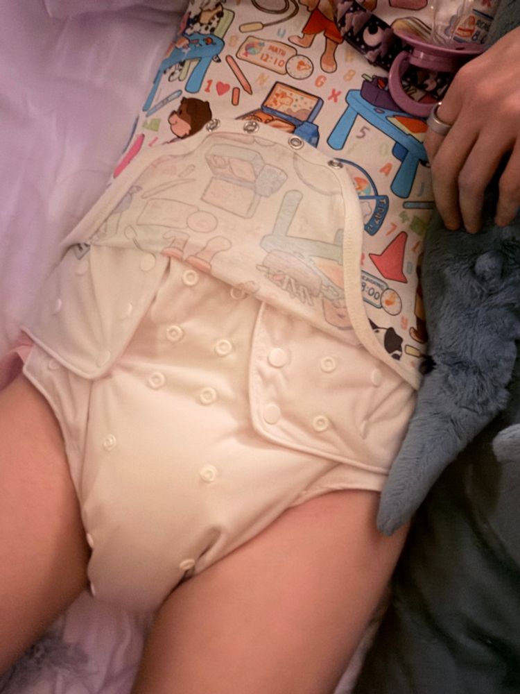 a woman lying in bed, wearing a onesie with its flap currently open, exposing a white diaper cover beneath (the diaper underneath is pink but you can’t really see it)