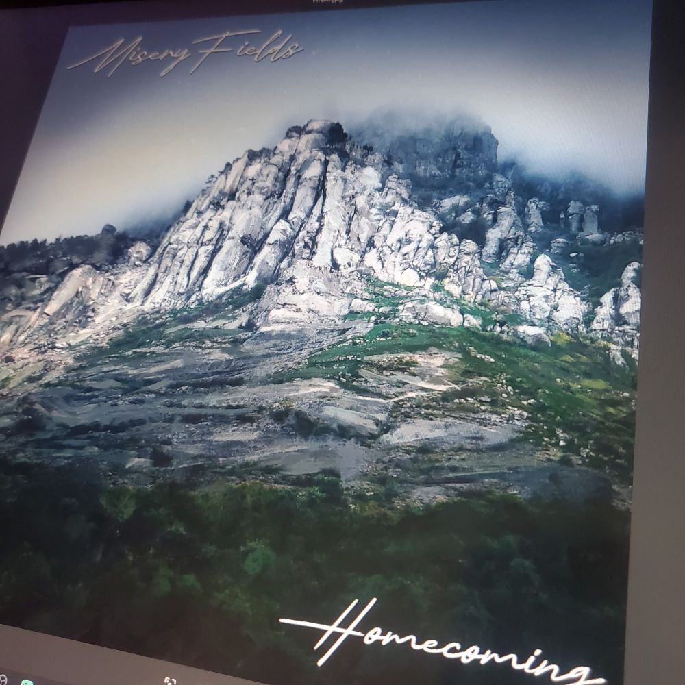 Photo of a screen showing an album cover, an image portraying a mountain covered in fog, the sky completely clouded. In the top left, text says MiseryFields. In the bottom right, text says Homecoming.