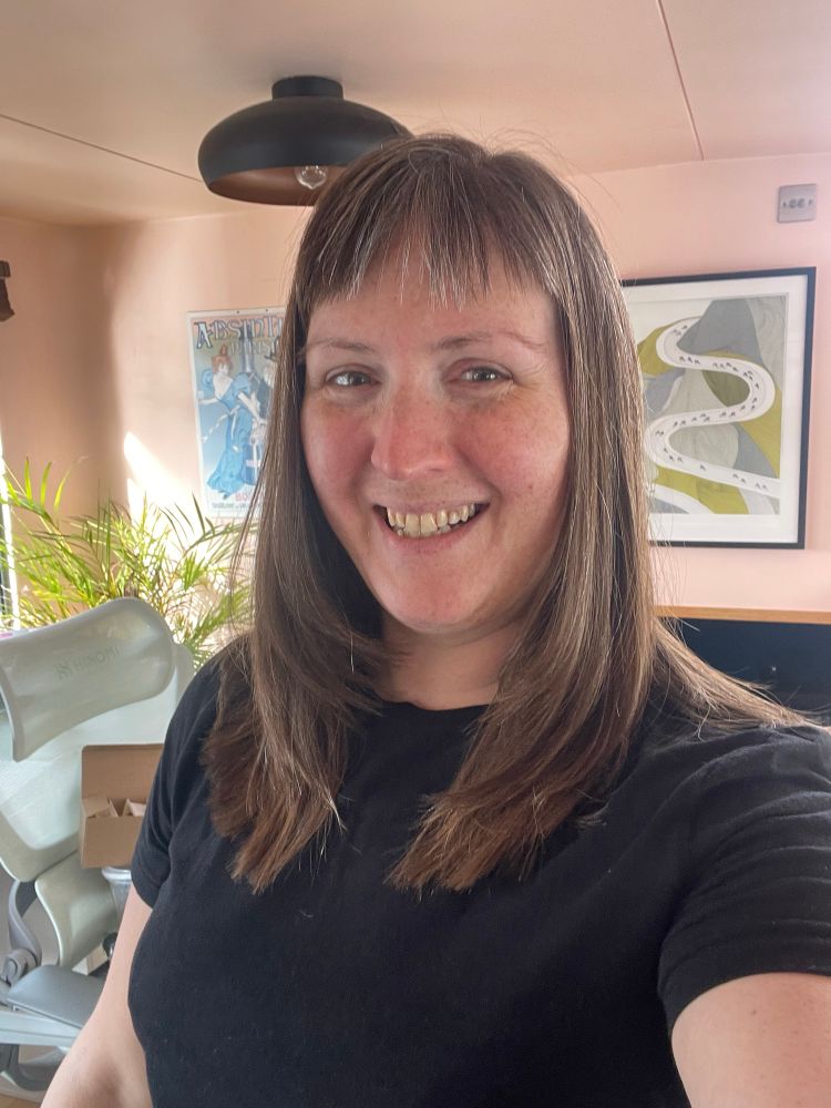 A white woman with straight brown hair is talking a selfie and smiling. Her hair is now only just past her shoulders and has nice layers framing her face.