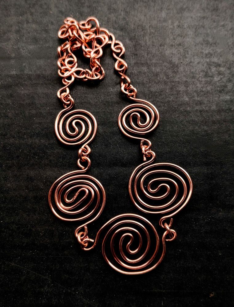 Sur un fond en carton ondulé noir, qui a un aspect velouté, un collier en cuivre 1,6 mm composé de trois grandes spirales centrales, puis une plus petite de chaque côté, et ensuite de la chaîne avec des maillons en forme d'infini. Le collier prend agréablement la lumière sur la surface des spirales. 