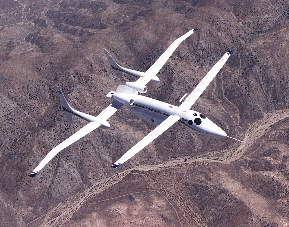 Scaled Composites Proteus in flight over a brown hilly landscape. It is a very weird-looking aircraft. Instead of a single windshield there are multiple circular windows around the cockpit, just aft of that there is a long slender wing, and at the very tail end of it there is another long slender wing, about a third longer than the front one. The engines are mounted in pods attached at the rear of the fuselage as well, and two vertical stabilisers are attached to the rear wing by booms that attach to it about a quarter of its span out from the fuselage.