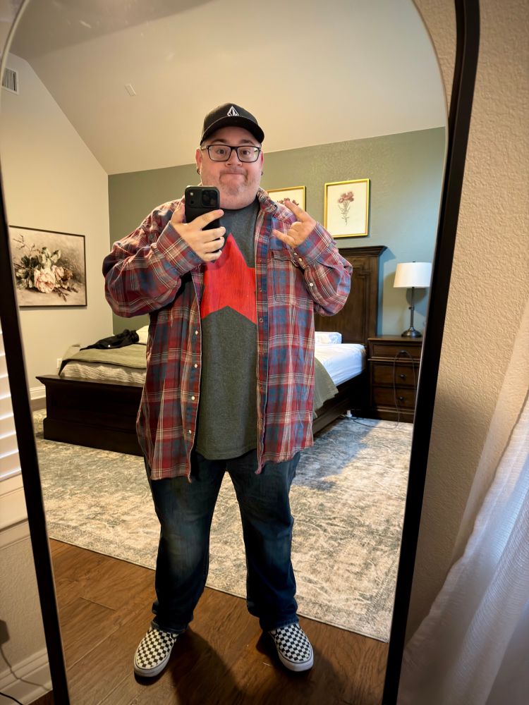 Me taking a selfie in front of a mirror. I’m wearing a graphic tee, flannel shirt, jeans, checkered Vans and a Volcom baseball hat. 