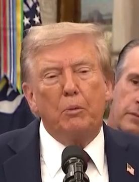 An ill orange man stands behind a microphone. He has puffy eyes and prominent swelling on his cheekbones