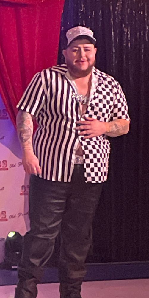 Drag king in black pants and a black and white checkered shirt and cap looking lovingly at their spouse off stage 