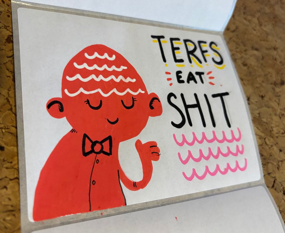 A pleasant red figure with a bow tie and maybe a tea cosy on their head smiling and holding one hand up next to the text “TERFS EAT SHIT”