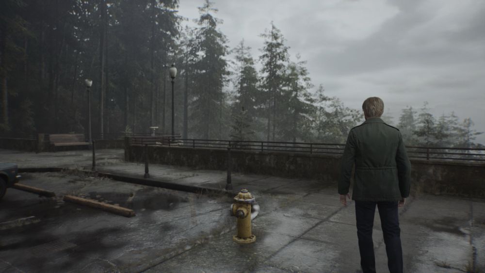 A screenshot of Silent Hill 2 Remake - a figure is stood at a rest stop overlooking a foggy pine forest 