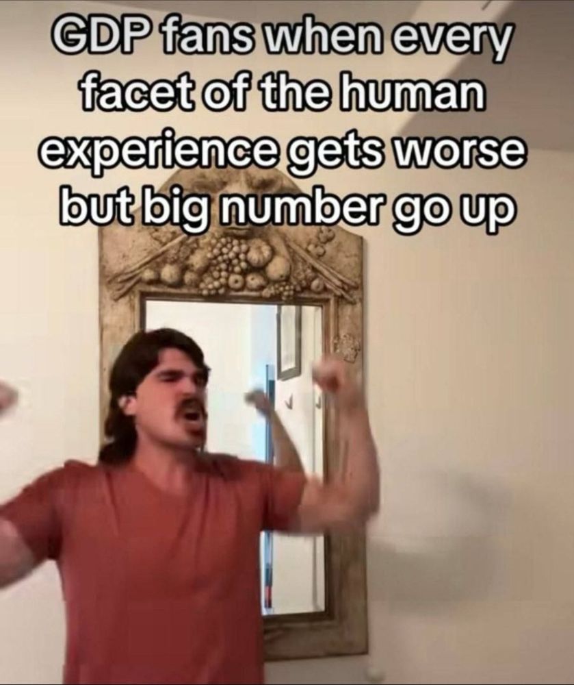 A person in a red shirt with medium long hair and a cool moustache is celebrating with the text “GDP fans when every facet of the human experience gets worse but big number go up”