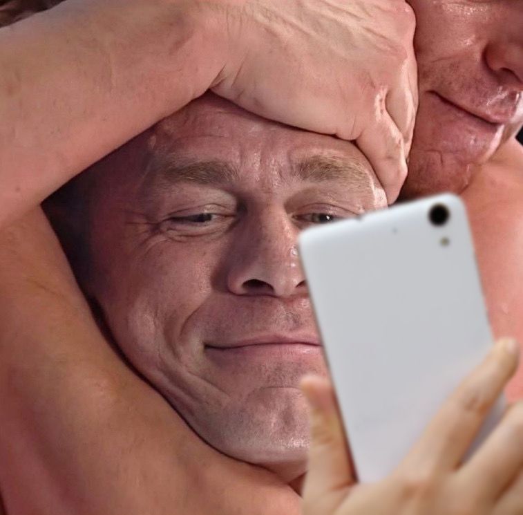 A tight close up of a man’s head being serenely squeezed in some burly arms as he looks ahead at his phone. 