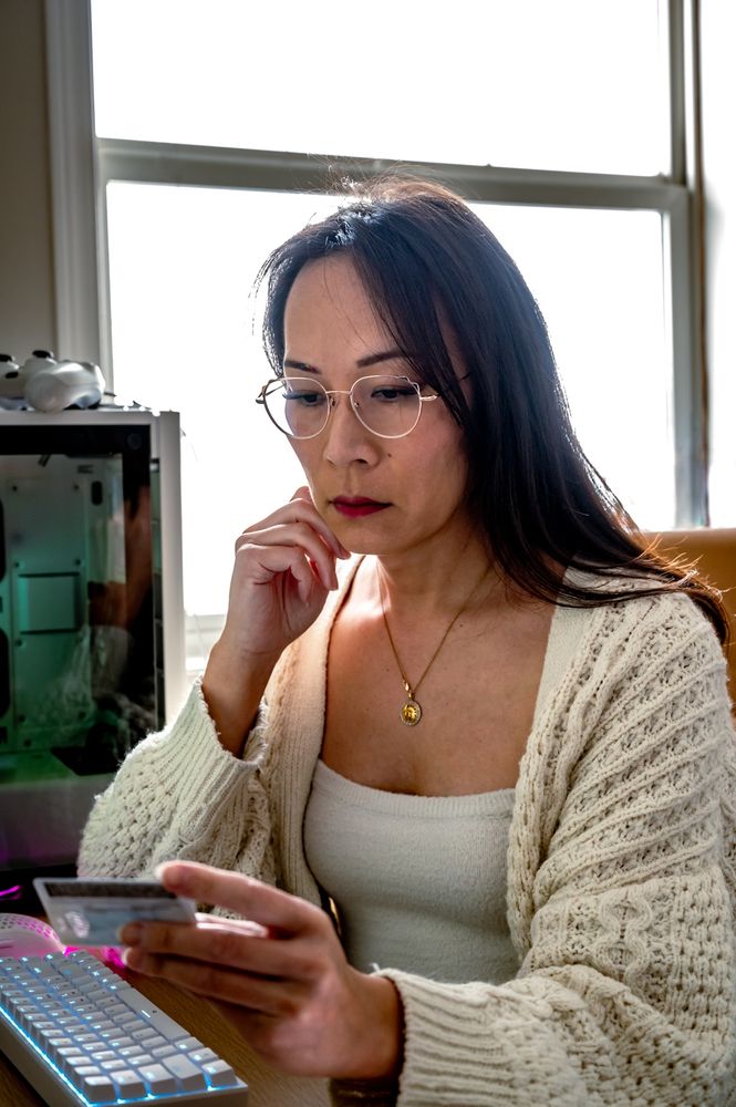 Cece has long black hair and gold rimmed glasses. She's wearing a slouchy chunky knit Cardigan over a knit cropped top. She's sitting beside her computer holding a driver's license and looking down at it pensively