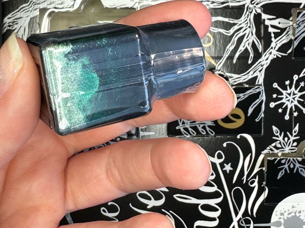 A caucasian hand holding the small Diamine Inkvent ink bottle over the Diamine Inkvent packaging to show the chameleon shimmer in the bottle. It’s greeny, blue.