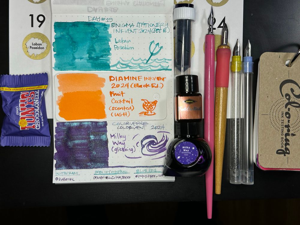Alt Text (as written):
A dotted notepad held open with a Traveler’s Company brand brass clip. The main notepad has three swatched inks, (will be described in detail momentarily) with those ink bottles to the right of the notepad, along with the tools that were used to make the swatches (L-R Tachikawa wooden nib holder with pink silicone grip holding a tubular steel Kakimori nib; a clear with silver sparkles proprietary Sailor Hocoro Dipton nib holder with a yellow Fude nib; a clear standard Sailor Hocoro with the blue fine nib). 

Note: Today included a scented ink, so the second swatch is on a separate Col-o-ring Ink Testing Card (shown to the right) from @wellappointeddesk.bsky.social that is done with a cotton swab & a Leonardt 30 pointed pen nib in an unnamed pink painted wooden holder.

To the left is a wrapped (tiny) Tony’s Chocolonely chocolate: navy wrapped “milk chocolate pretzel toffee”.

Everything is sitting on the Enigma Stationery Inkvent scratch off sheet that tells what the daily ink sample is. Day 19 is scratched off and says “Laban Poseidon”

The ink swatches, top to bottom are Enigma Stationery Inkvent 2024 set B Day 19 Laban Poseidon, which is a light to mid depth aqua-blue ink that has some shading in the main swatch. The ink was dry but wet at the same time. It didn’t stick to any nibs. The doodle is of some waves with a trident.

Middle ink swatch is Diamine Inkvent 2024 Black Edition, Fruit Cocktail (scented) a flat light to mid orange (like Irn Bru or Tang) with no shading or sheen. It smells *bad* like fruity hairpsray. The doodle is of a dessert glass & some fruit.

Bottom Ink is Colorverse Colorvent 2024 Milky Way (glistening) a deep plummy purple with fine, consistent aqua shimmer that echoes the Laban Poseidon. The doodle is a bad from-memory drawing of a galaxy.

The bottom of the page has social media info, titles in Poseidon & the names in Milky Way:
Instagram: @subgirl; Open IG / Inkyblog: @subgirl_inkygeek; BlueSky: @inkygeek.com