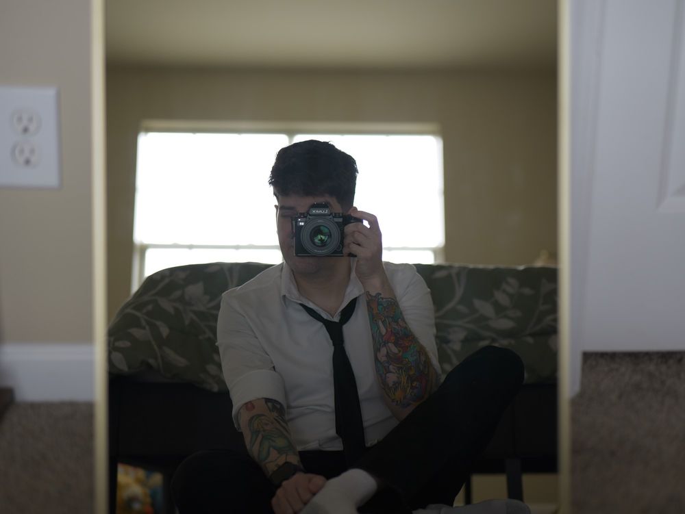 Self portrait of a ban dressed in a white shirt, black tie. Adorned with tattoos on his right and left forearms. Sitting in front of a mirror on the floor.