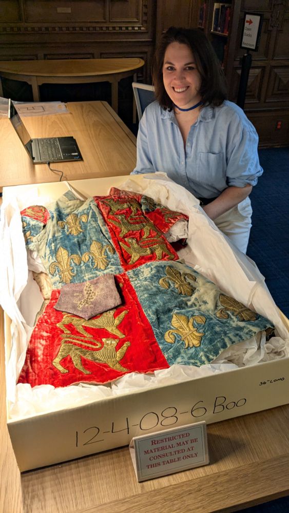 A red and blue velvet tunic with gold embroidered lion and fleur-de-lis sits in a box. A white woman with brown hair sits behind it, smiling. She is wearing a light blue button up shirt.