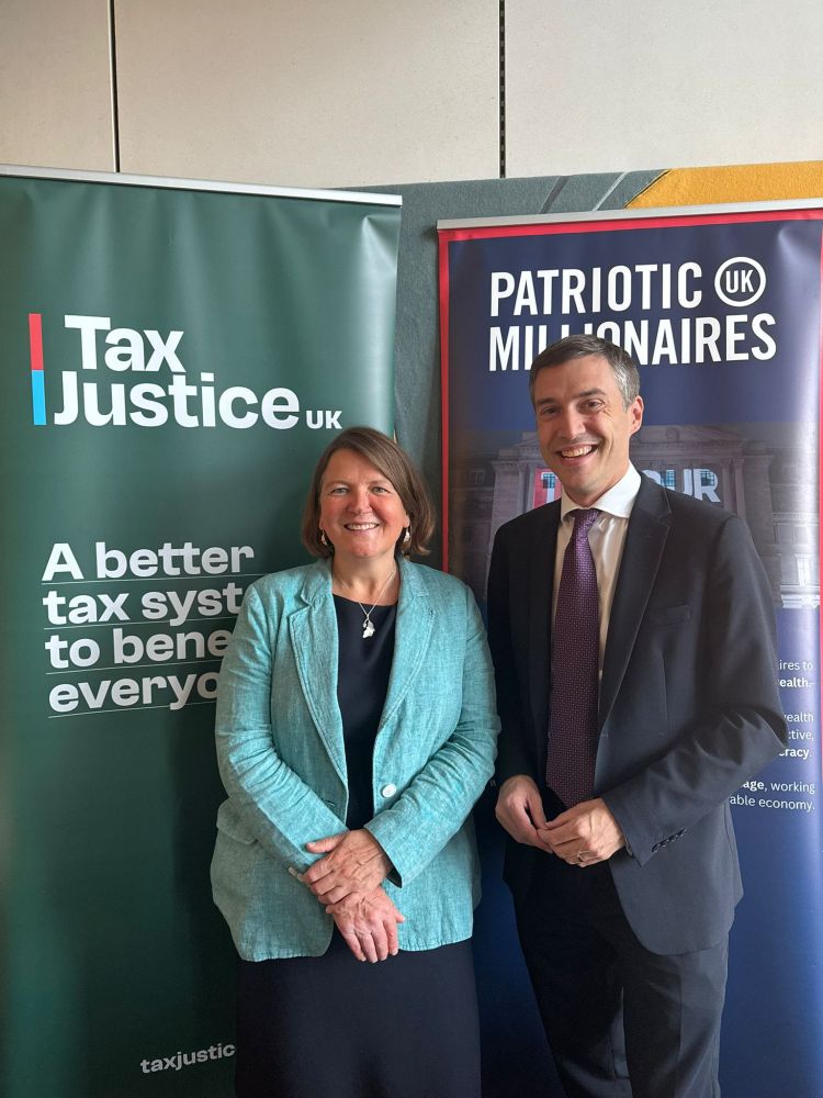 Ellie and Adrian Ramsay MP smiling in front of Patriotic Millionaires UK and Tax Justice UK banners