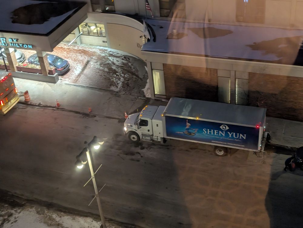 The shen Yun bus and truck parked in front of a double tree 