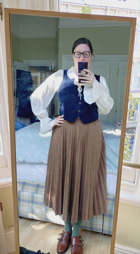 It is I, doing a mirror selfie, in a long brown pleated skirt, blue waistcoat and roguish cream-coloured blouse with frilly sleeve cuffs. It's gently implying fantasy librarian or light academia.