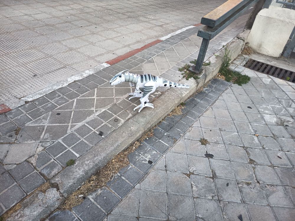 Juguete de ¿robot dinosaurio? ¿dinosaurio robot? en mitad de la acera. 
