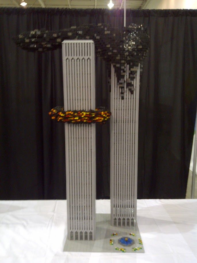 A Lego rendtion of the Twin Towers in NYC made of gray plastic bricks depicting them after the impact of the second plane on 09/11/2001; tower on the left has an explosion depicted about 2/3rd of the way up while the tower on the right has a plume of smoke drifting up and to the left behind the tower on the left