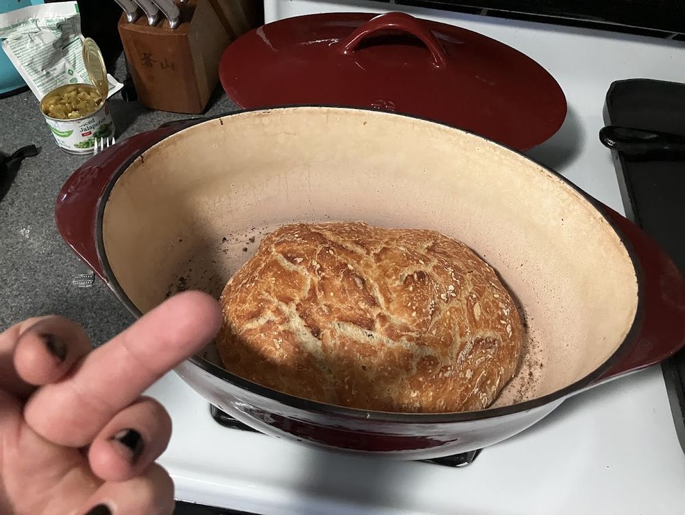 A brown loaf of baked sourdough bread in a burgundy Dutch oven on top of a white stove, lid off on the back burner, my left hand in the foreground with middle finger extended at said loaf